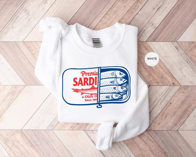 May include: White sweatshirt featuring a graphic of a sardine can with the words "Premium Sardines in Olive Oil Since 1900". The sardine can design is in red and blue. The sweatshirt is white.