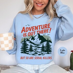 K&ouml;nnte beinhalten: Hellblaues Sweatshirt mit dem Text "ADVENTURE IS OUT THERE!" in Rot, &uuml;ber einer Berg- und Waldgrafik. Der Text "BUT SO ARE SERIAL KILLERS" steht darunter. Das Sweatshirt ist hellblau.