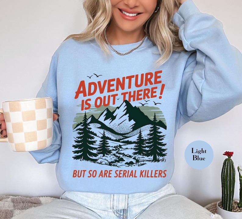 K&ouml;nnte beinhalten: Hellblaues Sweatshirt mit dem Text "ADVENTURE IS OUT THERE!" in Rot, &uuml;ber einer Berg- und Waldgrafik. Der Text "BUT SO ARE SERIAL KILLERS" steht darunter. Das Sweatshirt ist hellblau.
