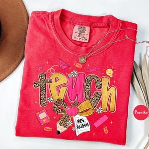 May include: Red Comfort Colors t-shirt with a teacher-themed graphic. The word "teach" is displayed in a mix of leopard print and gold lettering, with school-related illustrations. A gold necklace is draped across the shirt.
