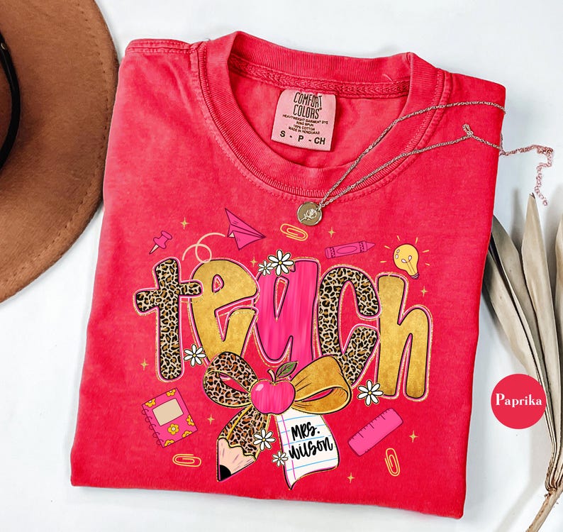 May include: Red Comfort Colors t-shirt with a teacher-themed graphic. The word "teach" is displayed in a mix of leopard print and gold lettering, with school-related illustrations. A gold necklace is draped across the shirt.
