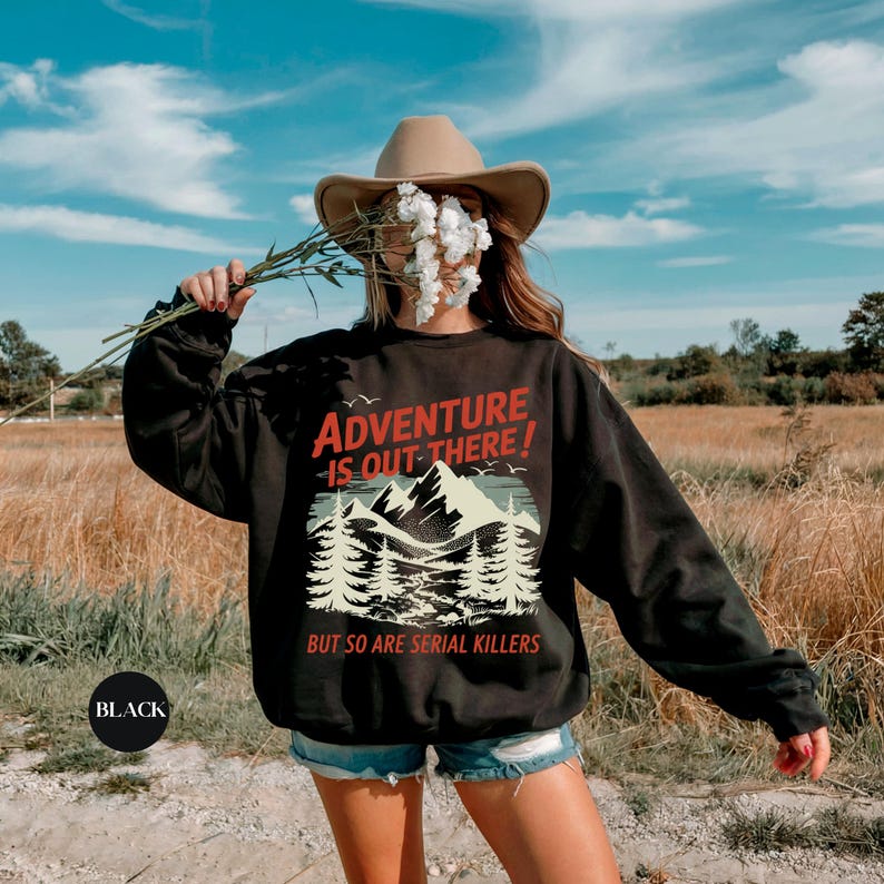K&ouml;nnte beinhalten: Schwarzes Sweatshirt mit dem Text "ADVENTURE IS OUT THERE! BUT SO ARE SERIAL KILLERS" in Rot und Wei&szlig;. Die Grafik zeigt eine Bergkette und B&auml;ume. Das Sweatshirt wird mit Jeansshorts getragen.