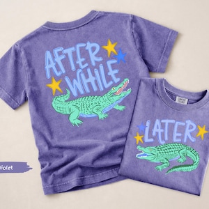 Puede incluir: Dos camisetas violetas con gr&aacute;ficos de caimanes. Una camiseta dice "AFTER WHILE" con un caim&aacute;n verde y estrellas amarillas y azules. La otra camiseta dice "LATER" con un caim&aacute;n verde y estrellas amarillas y azules.