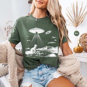 Może przedstawiać: Zielona koszulka z białym nadrukiem przedstawiającym kota patrzącego w g&oacute;rę na UFO. UFO emituje wiązki światła. Nadruk zawiera r&oacute;wnież małą rybę latającą w powietrzu.