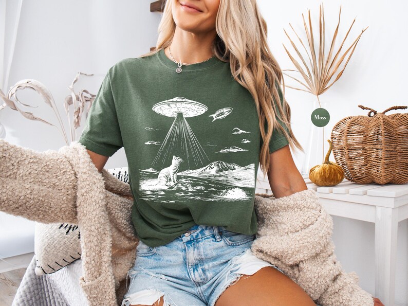 Może przedstawiać: Zielona koszulka z białym nadrukiem przedstawiającym kota patrzącego w g&oacute;rę na UFO. UFO emituje wiązki światła. Nadruk zawiera r&oacute;wnież małą rybę latającą w powietrzu.