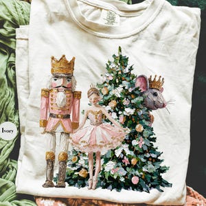 May include: Ivory-colored t-shirt featuring a whimsical illustration of a nutcracker, ballerina, mouse king, and Christmas tree. The design includes gold and pink accents, creating a festive and artistic aesthetic. The shirt is made of a soft material.
