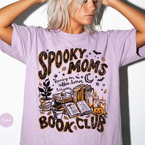 May include: A light purple short-sleeve t-shirt featuring a graphic print with the text "SPOOKY MOMS BOOK CLUB" and "Running on coffee, horror & chapters". The design includes illustrations of stacked books, a skull, a pumpkin, candles, and plants.