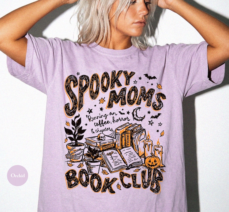 May include: A light purple short-sleeve t-shirt featuring a graphic print with the text "SPOOKY MOMS BOOK CLUB" and "Running on coffee, horror & chapters". The design includes illustrations of stacked books, a skull, a pumpkin, candles, and plants.