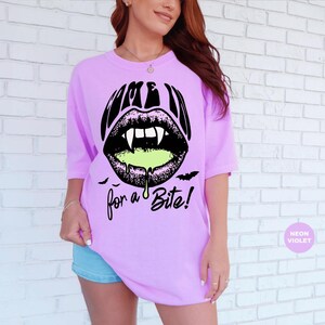 May include: A neon violet t-shirt featuring a black graphic of lips with vampire fangs and dripping lime green liquid. The text reads "COME IN for a Bite!" in a stylized black font, accompanied by small black bat silhouettes below the lips.