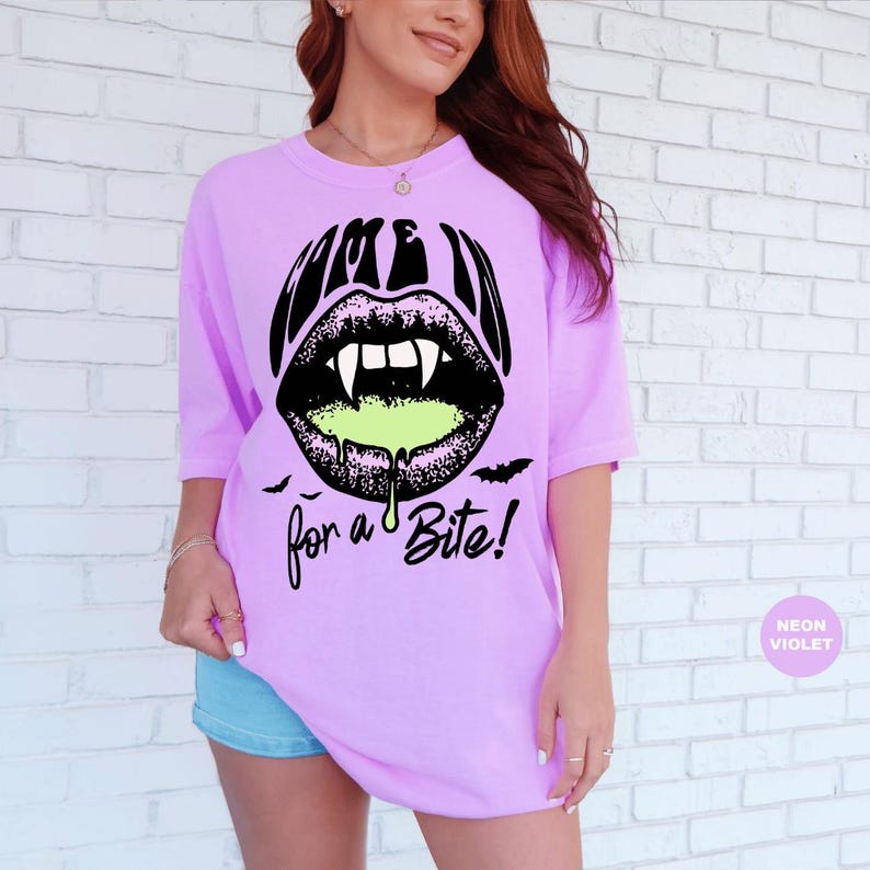 May include: A neon violet t-shirt featuring a black graphic of lips with vampire fangs and dripping lime green liquid. The text reads "COME IN for a Bite!" in a stylized black font, accompanied by small black bat silhouettes below the lips.