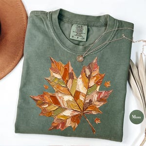 Può includere: Una t-shirt verde muschio con una grande grafica di una foglia d'acero dipinta con una texture in stile impasto nei toni dell'arancione, marrone, giallo e rosso. La maglietta ha un girocollo e un'etichetta Comfort Colors stampata all'interno.