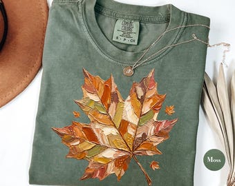 May include: A moss green t-shirt featuring a large, textured, impasto-style painted maple leaf graphic in shades of orange, brown, yellow, and red. The shirt has a crew neckline and a Comfort Colors brand label printed on the inside neck area.