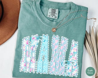 May include: A light green short-sleeve t-shirt with a crew neckline and a Comfort Colors label. The front features the word MAMA in large letters, decorated with floral and polka dot patterns, white lace trim, and two small light blue ribbons.