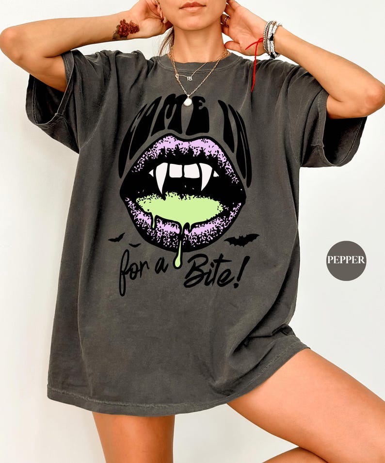May include: A charcoal gray short-sleeve t-shirt featuring a graphic of purple lips with white vampire fangs and green dripping liquid. The text reads "COME IN" above the lips and "for a Bite!" below, accompanied by two small black bat silhouettes.