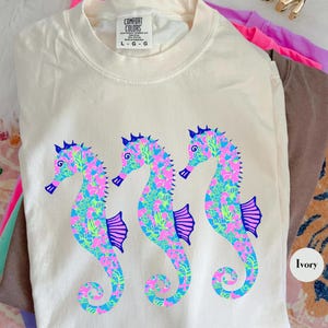 May include: Ivory-colored Comfort Colors t-shirt featuring three seahorse designs. The seahorses are decorated with a colorful pattern of pink, blue, and green. The shirt has a crew neck and short sleeves. The tag reads L-G-G.