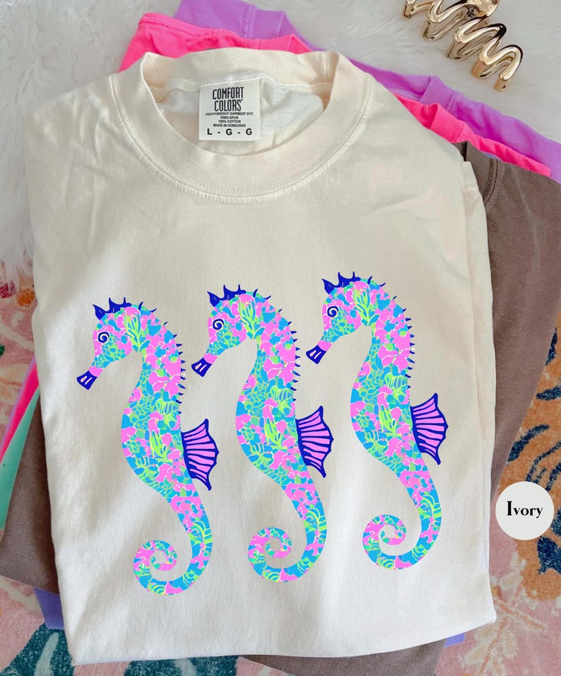 May include: Ivory-colored Comfort Colors t-shirt featuring three seahorse designs. The seahorses are decorated with a colorful pattern of pink, blue, and green. The shirt has a crew neck and short sleeves. The tag reads L-G-G.