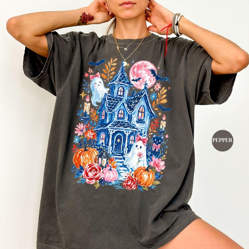 K&ouml;nnte beinhalten: Ein anthrazitfarbenes Kurzarm-T-Shirt mit einem farbenfrohen Grafikdruck eines blauen Spukhauses, zwei wei&szlig;en Geistern mit rosa Schleifen, orangefarbenen K&uuml;rbissen, roten Rosen, einer schwarzen Katze und einem gro&szlig;en rosa Mond mit Flederm&auml;usen.