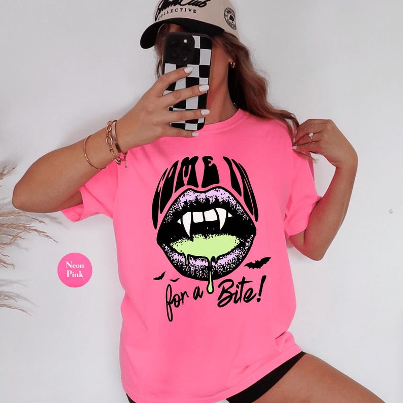 May include: A neon pink t-shirt featuring a black graphic of vampire lips with white fangs and dripping lime green liquid. The text above the lips reads "COME IN" and the text below reads "for a Bite!" with small black bats on the sides.