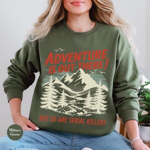 K&ouml;nnte beinhalten: Milit&auml;rgr&uuml;nes Sweatshirt mit dem Text "ADVENTURE IS OUT THERE! BUT SO ARE SERIAL KILLERS" in Rot und Wei&szlig;. Die Grafik zeigt eine Bergkette, B&auml;ume und V&ouml;gel.