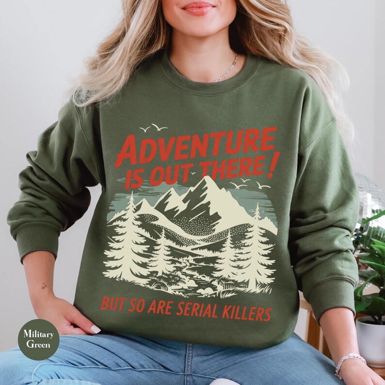 K&ouml;nnte beinhalten: Milit&auml;rgr&uuml;nes Sweatshirt mit dem Text "ADVENTURE IS OUT THERE! BUT SO ARE SERIAL KILLERS" in Rot und Wei&szlig;. Die Grafik zeigt eine Bergkette, B&auml;ume und V&ouml;gel.