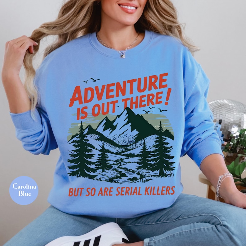 K&ouml;nnte beinhalten: Carolina-blaues Sweatshirt mit dem Text "ADVENTURE IS OUT THERE! BUT SO ARE SERIAL KILLERS" in Rot und Wei&szlig;. Die Grafik zeigt eine Bergkette, Kiefern und V&ouml;gel. Das Sweatshirt hat einen Rundhalsausschnitt.