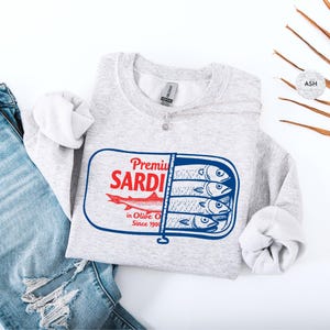 May include: A light gray sweatshirt with a graphic of a sardine can. The can design features the words "Premium Sardi" in red, a red fish, and the text "in Olive Oil Since 1900". The sweatshirt is paired with blue jeans.