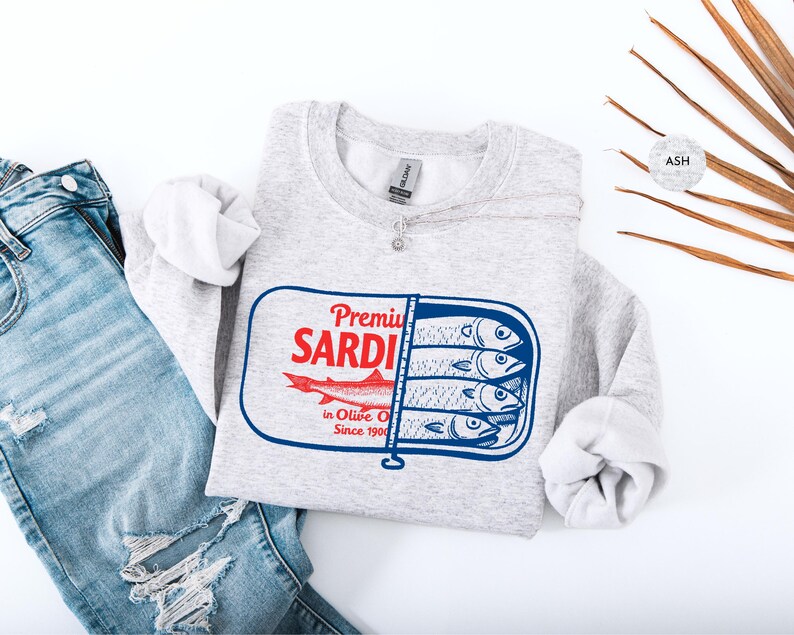 May include: A light gray sweatshirt with a graphic of a sardine can. The can design features the words "Premium Sardi" in red, a red fish, and the text "in Olive Oil Since 1900". The sweatshirt is paired with blue jeans.