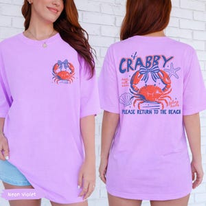 Puede incluir: Camiseta violeta ne&oacute;n con un gr&aacute;fico de cangrejo en la parte delantera y trasera. La parte delantera presenta un cangrejo rojo con un lazo azul. La parte trasera tiene las palabras "If Crabby" encima del cangrejo y "Please Return to the Beach" debajo.