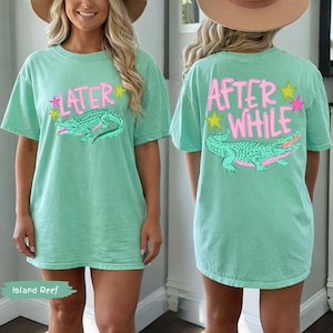 May include: A mint green t-shirt with a graphic of an alligator and the text "LATER" on the front. The back of the shirt features the text "AFTER WHILE" and a matching alligator graphic. The shirt has a relaxed fit.