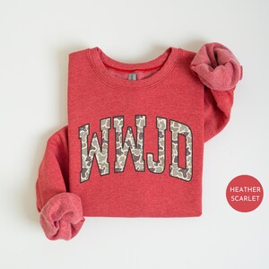 May include: A heather scarlet red sweatshirt with the letters "WWJD" in a camouflage pattern across the chest. The sweatshirt has a crew neck and long sleeves with rolled-up cuffs. A small, round sticker with the text "HEATHER SCARLET" is in the bottom right corner.