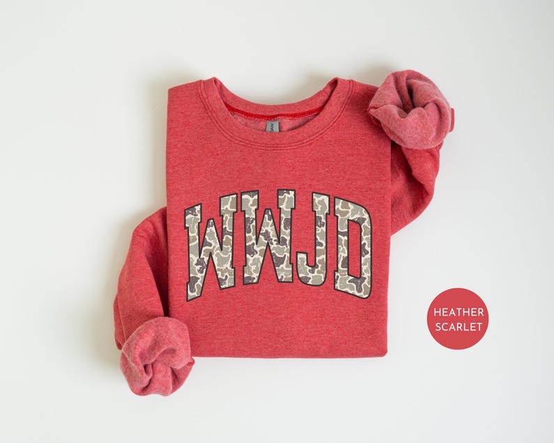 May include: A heather scarlet red sweatshirt with the letters "WWJD" in a camouflage pattern across the chest. The sweatshirt has a crew neck and long sleeves with rolled-up cuffs. A small, round sticker with the text "HEATHER SCARLET" is in the bottom right corner.