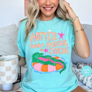 Puede incluir: Camiseta azul claro con el texto "Hotter Than a Hoochie Coochie" en rosa y naranja. La camiseta presenta un cocodrilo de dibujos animados flotando en una boya naranja y rosa. La camiseta es de color Chalky Mint.