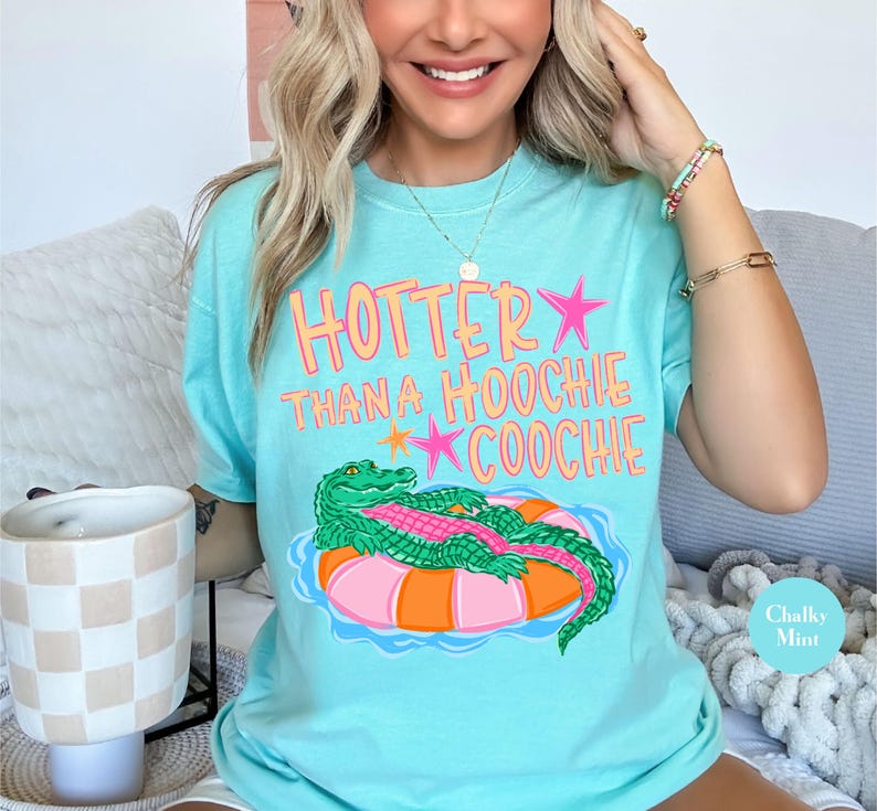 Puede incluir: Camiseta azul claro con el texto "Hotter Than a Hoochie Coochie" en rosa y naranja. La camiseta presenta un cocodrilo de dibujos animados flotando en una boya naranja y rosa. La camiseta es de color Chalky Mint.