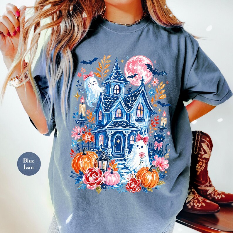 K&ouml;nnte beinhalten: Ein blau-graues Kurzarm-T-Shirt mit einem farbenfrohen Grafikdruck eines Spukhauses, Geistern mit rosa Schleifen, orangefarbenen K&uuml;rbissen, roten Rosen, einem Vollmond, Flederm&auml;usen und brennenden Kerzen. Der Text Blue Jean steht in einem Kreis.