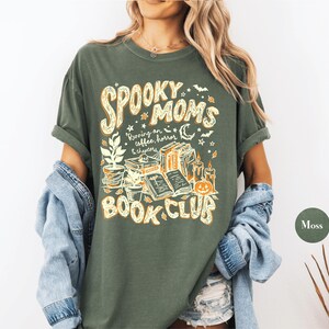 May include: A moss green short-sleeve t-shirt featuring a graphic print in cream and orange. The text reads "SPOOKY MOMS BOOK CLUB" and "Running on coffee, horror & chapters," accompanied by illustrations of books, potted plants, candles, and a pumpkin.