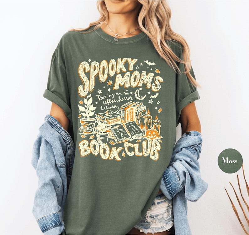 May include: A moss green short-sleeve t-shirt featuring a graphic print in cream and orange. The text reads "SPOOKY MOMS BOOK CLUB" and "Running on coffee, horror & chapters," accompanied by illustrations of books, potted plants, candles, and a pumpkin.