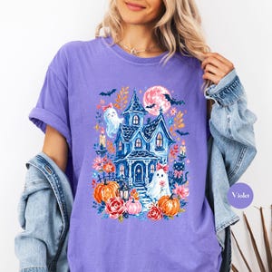 K&ouml;nnte beinhalten: Ein violettes T-Shirt mit einem Grafikdruck eines blauen Spukhauses, Vollmond, Flederm&auml;usen, zwei Geistern mit rosa Schleifen, orangefarbenen K&uuml;rbissen und roten Blumen. Das Design zeigt zudem brennende Kerzen und eine schwarze Katze.