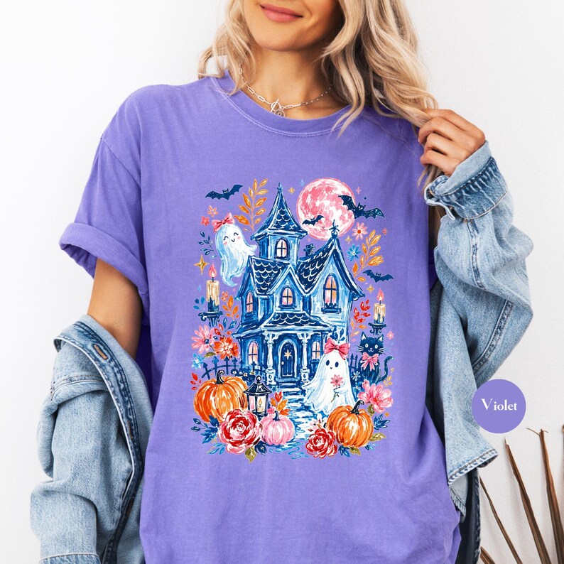 K&ouml;nnte beinhalten: Ein violettes T-Shirt mit einem Grafikdruck eines blauen Spukhauses, Vollmond, Flederm&auml;usen, zwei Geistern mit rosa Schleifen, orangefarbenen K&uuml;rbissen und roten Blumen. Das Design zeigt zudem brennende Kerzen und eine schwarze Katze.