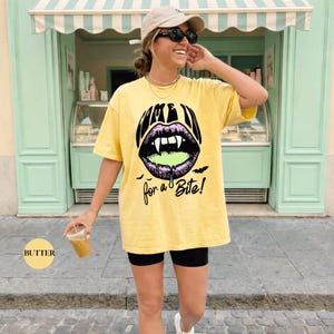 May include: A butter yellow short-sleeve t-shirt featuring a graphic of purple lips with white fangs and green dripping liquid. The text reads "COME IN for a Bite!" in black, accompanied by small black bat silhouettes below the lips.