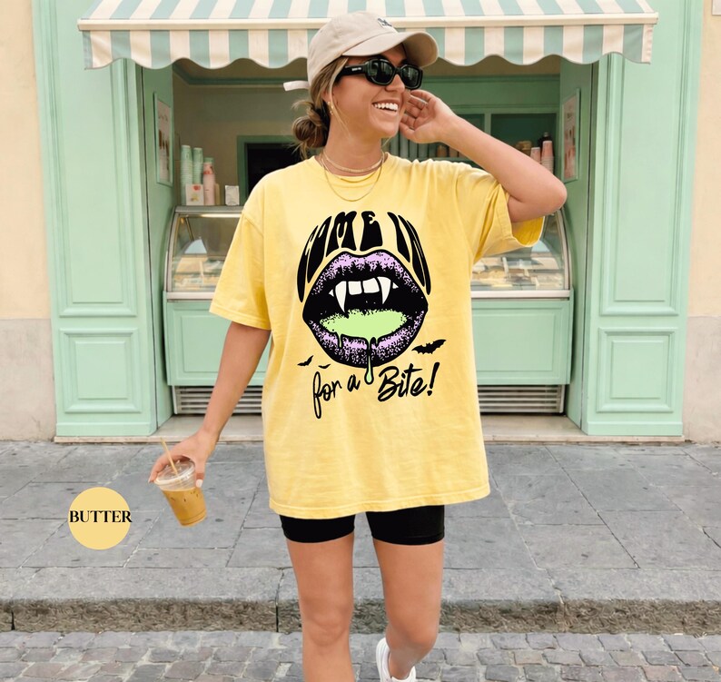 May include: A butter yellow short-sleeve t-shirt featuring a graphic of purple lips with white fangs and green dripping liquid. The text reads "COME IN for a Bite!" in black, accompanied by small black bat silhouettes below the lips.