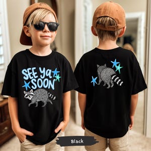 May include: Black t-shirt featuring a raccoon graphic and the text "See Ya Soon" in blue. The design includes blue and teal stars. The back of the shirt also has a raccoon graphic and stars. The shirt is a youth size.