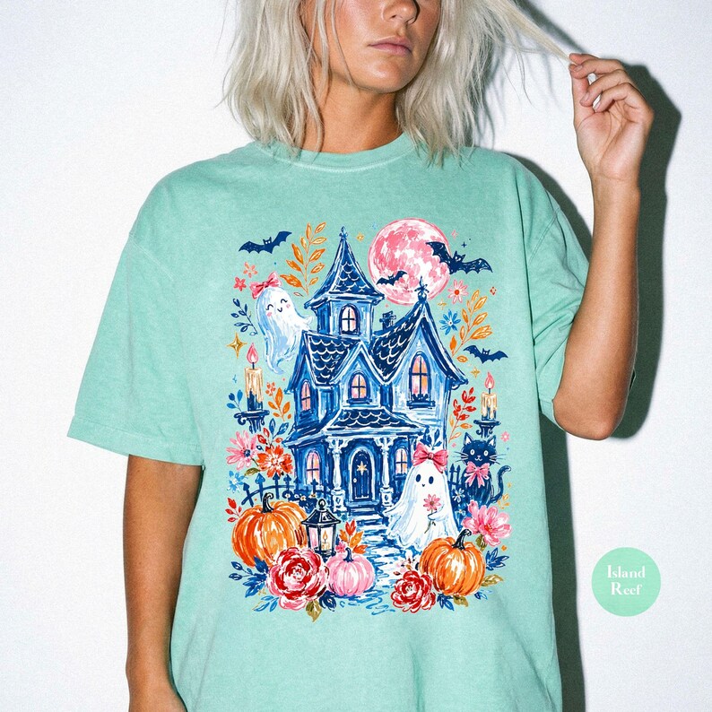 K&ouml;nnte beinhalten: Ein mintgr&uuml;nes T-Shirt mit einem farbenfrohen Grafikdruck eines blauen Spukhauses, zwei Geistern mit rosa Schleifen, orangefarbenen K&uuml;rbissen, einer schwarzen Katze, Flederm&auml;usen, einem gro&szlig;en rosa Mond und verschiedenen floralen Elementen.