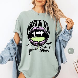 May include: A sage green short-sleeve t-shirt featuring a graphic of black lips with white fangs and dripping neon green liquid. The text above the lips reads COME IN and the text below reads for a Bite! with two small black bat silhouettes.