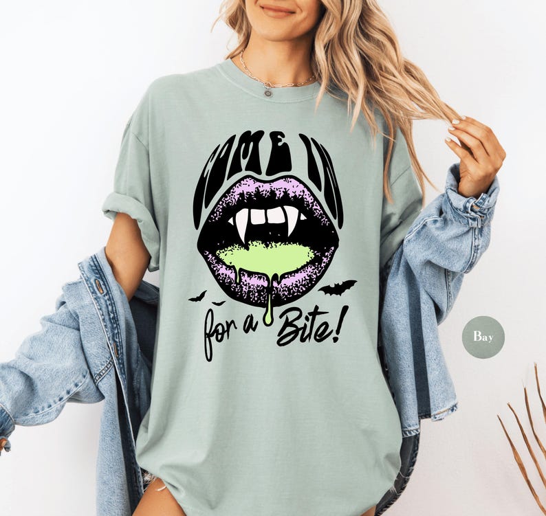 May include: A sage green short-sleeve t-shirt featuring a graphic of black lips with white fangs and dripping neon green liquid. The text above the lips reads COME IN and the text below reads for a Bite! with two small black bat silhouettes.