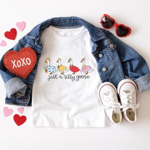 以下が含まれることがあります： 白いTシャツに、異なるコスチュームを着た4羽のガチョウと「Just a silly goose」という文字がプリントされています。Tシャツはデニムジャケット、赤いハート型のサングラス、白いスニーカーと合わせています。