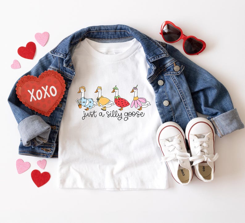 以下が含まれることがあります： 白いTシャツに、異なるコスチュームを着た4羽のガチョウと「Just a silly goose」という文字がプリントされています。Tシャツはデニムジャケット、赤いハート型のサングラス、白いスニーカーと合わせています。