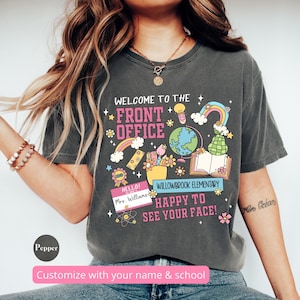 Op de afbeelding: Een donkergrijs T-shirt met een kleurrijk grafisch ontwerp met de woorden "Welkom bij het kantoor" en een verscheidenheid aan schoolthema-illustraties. De tekst "Willowbrook Elementary" en "Leuk je te zien!" zijn ook in het ontwerp opgenomen.