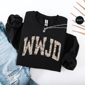 May include: Black crewneck sweatshirt with the letters WWJD in a camouflage pattern. A silver necklace with a circular pendant rests on the sweatshirt. The sweatshirt is paired with light blue ripped jeans.