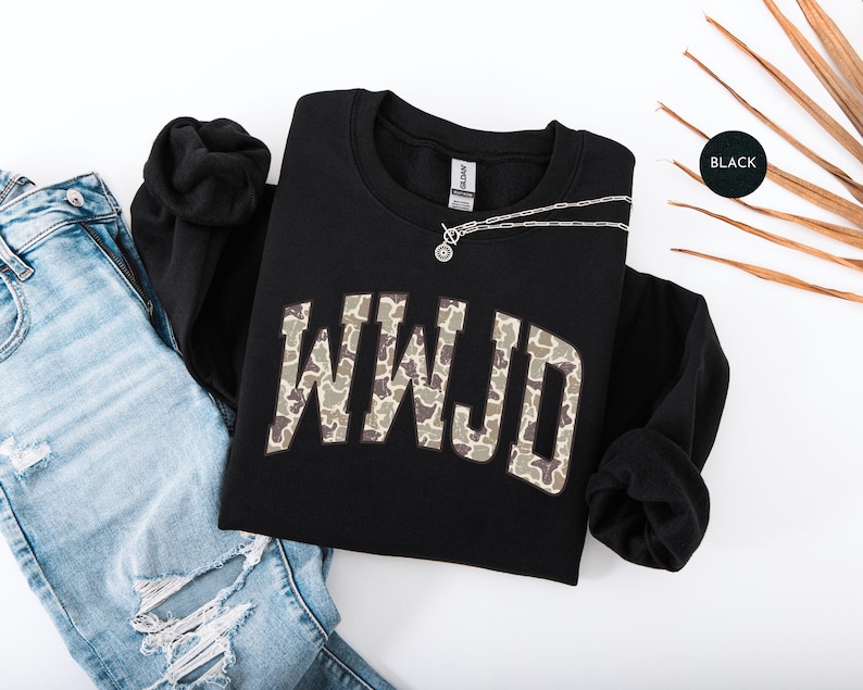 May include: Black crewneck sweatshirt with the letters WWJD in a camouflage pattern. A silver necklace with a circular pendant rests on the sweatshirt. The sweatshirt is paired with light blue ripped jeans.