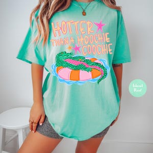 Puede incluir: Camiseta turquesa con el texto "Hotter Than a Hoochie Coochie" en rosa y naranja. La camiseta presenta un gr&aacute;fico de un caim&aacute;n verde sobre un flotador rosa y naranja. La camiseta tiene un ajuste holgado y c&oacute;modo.